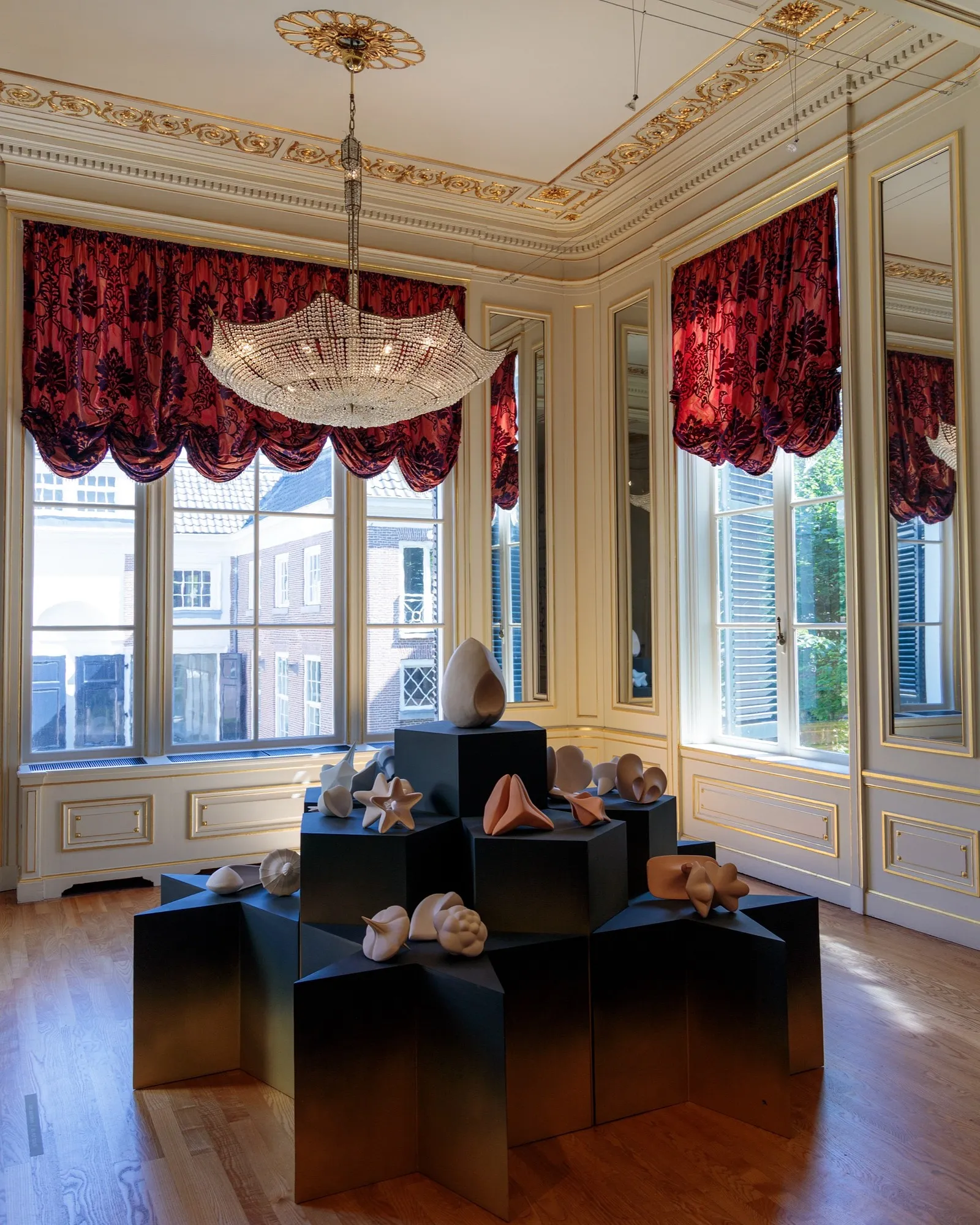 A gilded palace room with red damask curtains and an upturned umbrella-shaped crystal chandelier above pale sculptural forms on dark plinths.