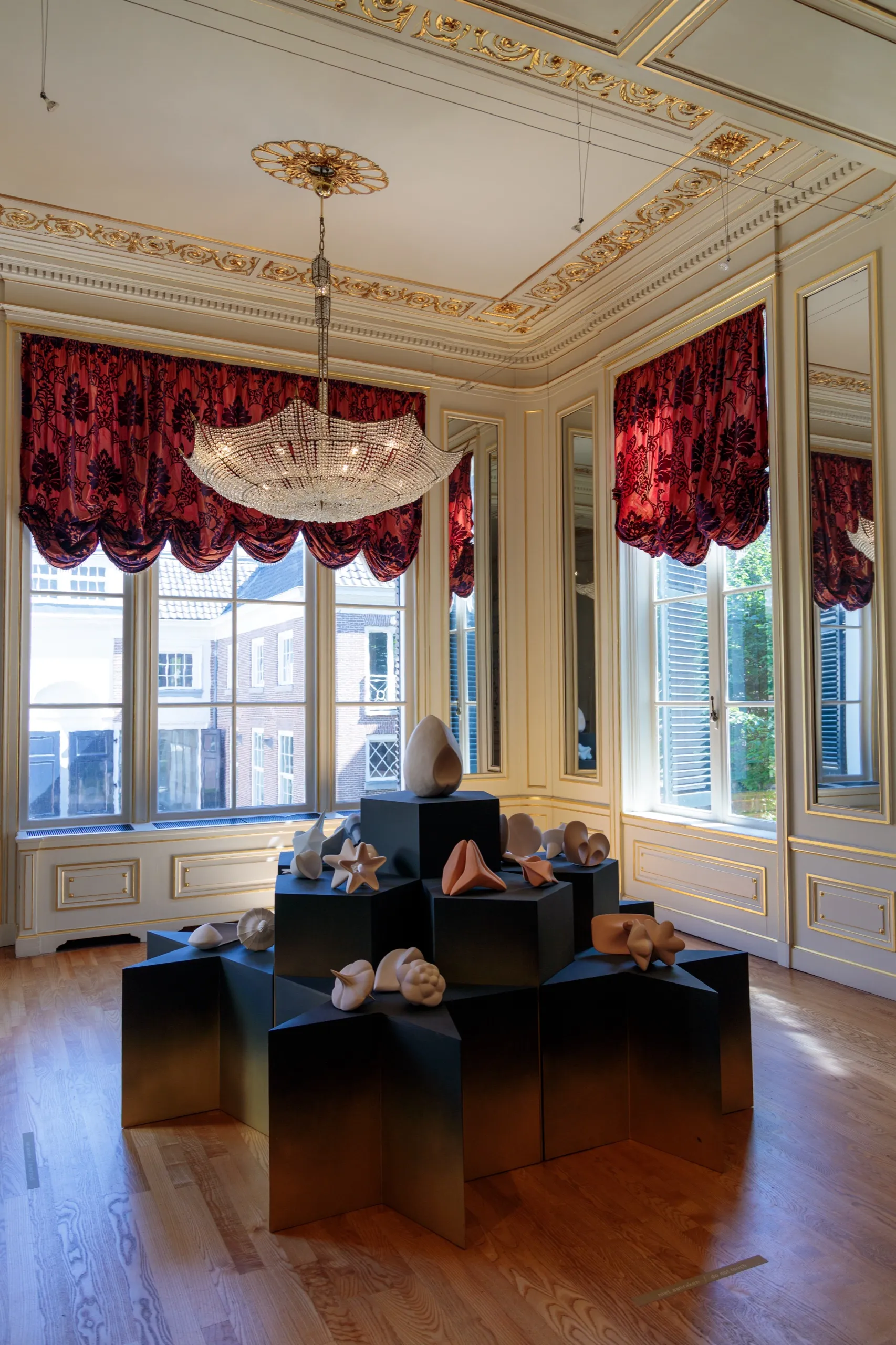 A gilded palace room with red damask curtains and an upturned umbrella-shaped crystal chandelier above pale sculptural forms on dark plinths.