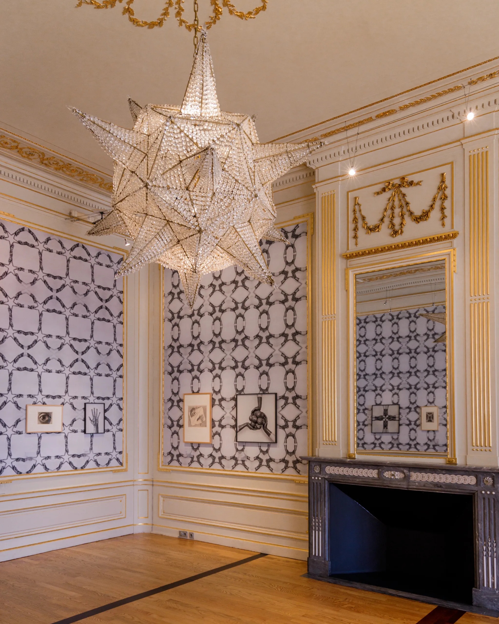 A large faceted crystal star chandelier in a gilded palace room, reflected in a tall mirror, with small framed Escher prints on the patterned walls below.