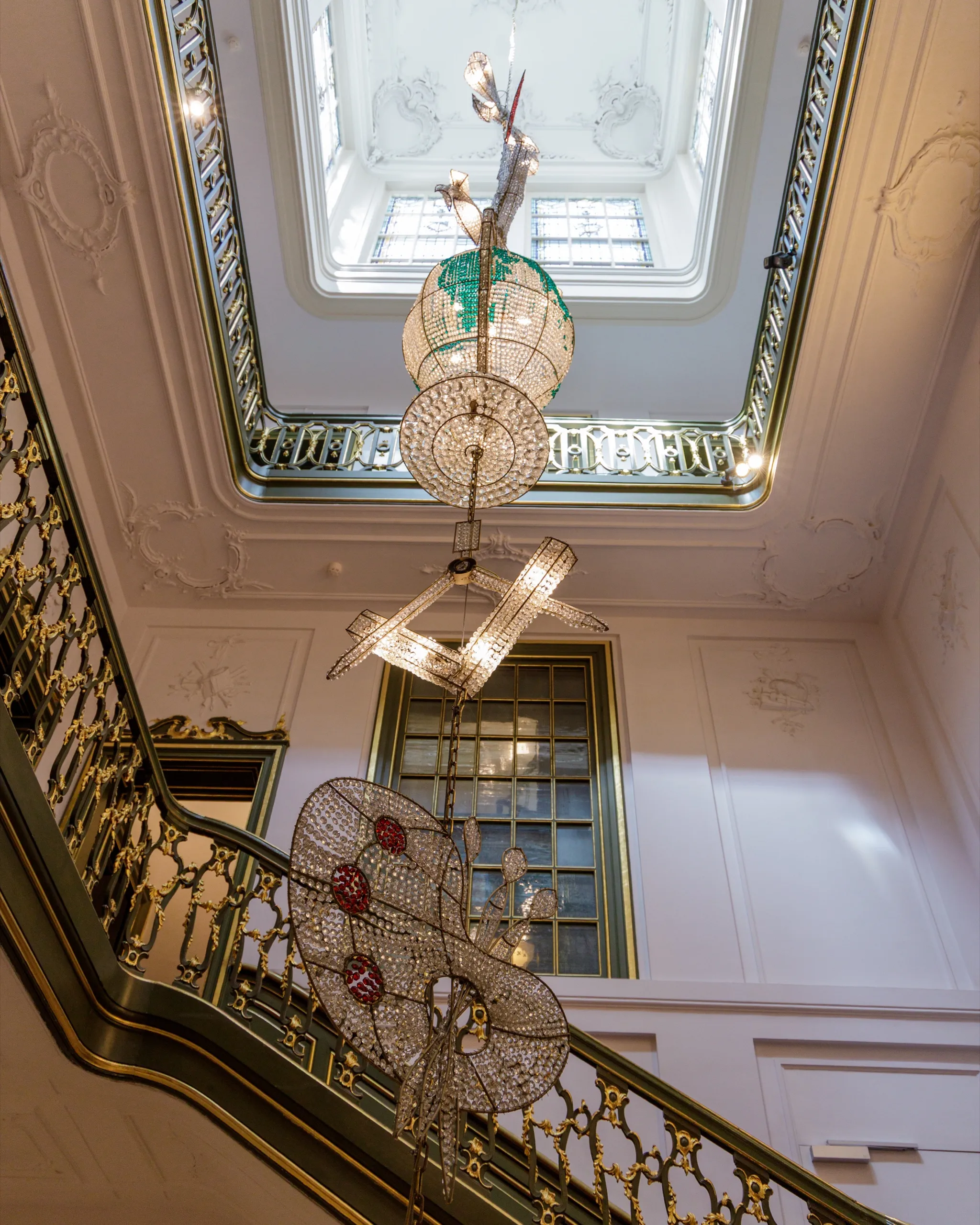 Looking up a grand palace stairwell at a stacked run of crystal chandeliers under an ornate skylight.
