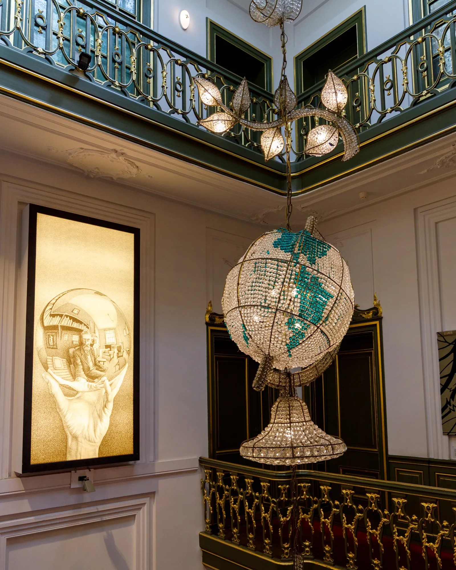 A large backlit reproduction of Escher's mirrored-sphere self-portrait glowing on a panelled stairwell wall, next to a crystal globe chandelier.