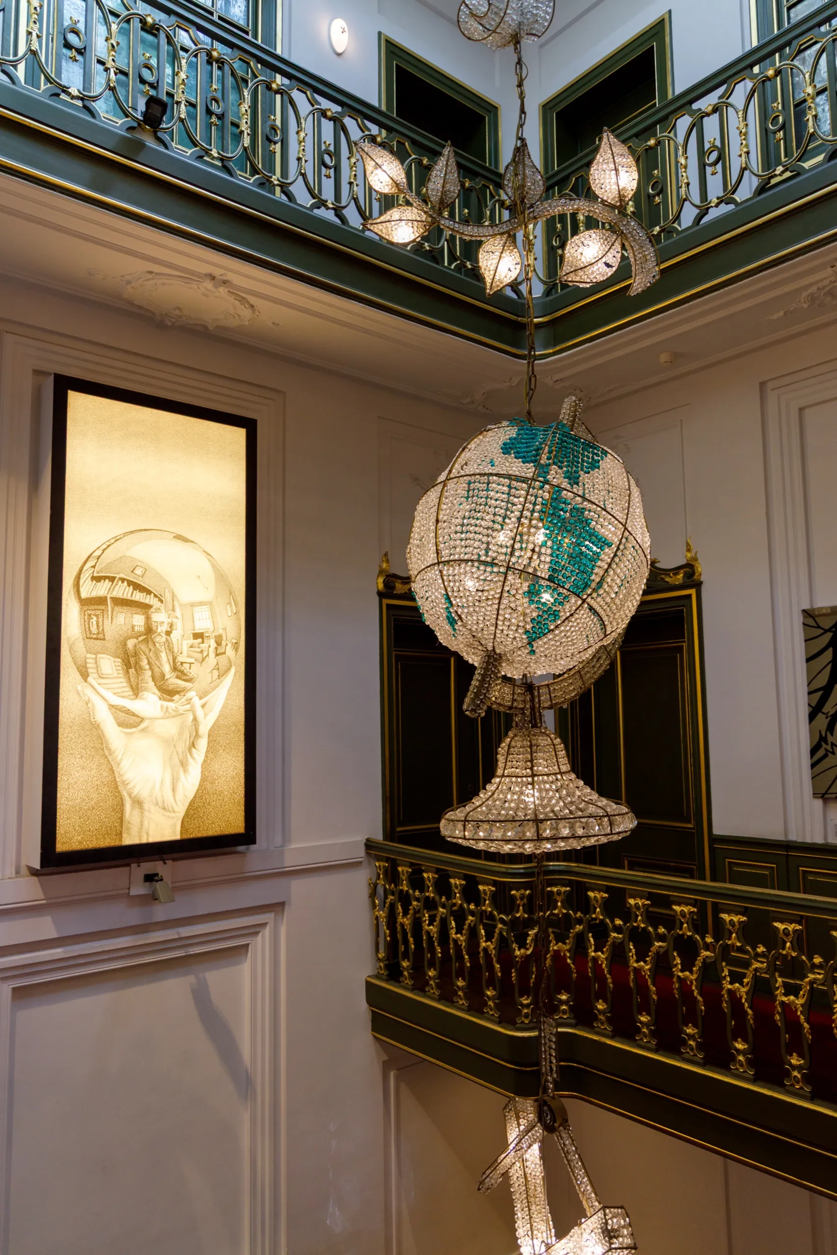 A large backlit reproduction of Escher's mirrored-sphere self-portrait glowing on a panelled stairwell wall, next to a crystal globe chandelier.