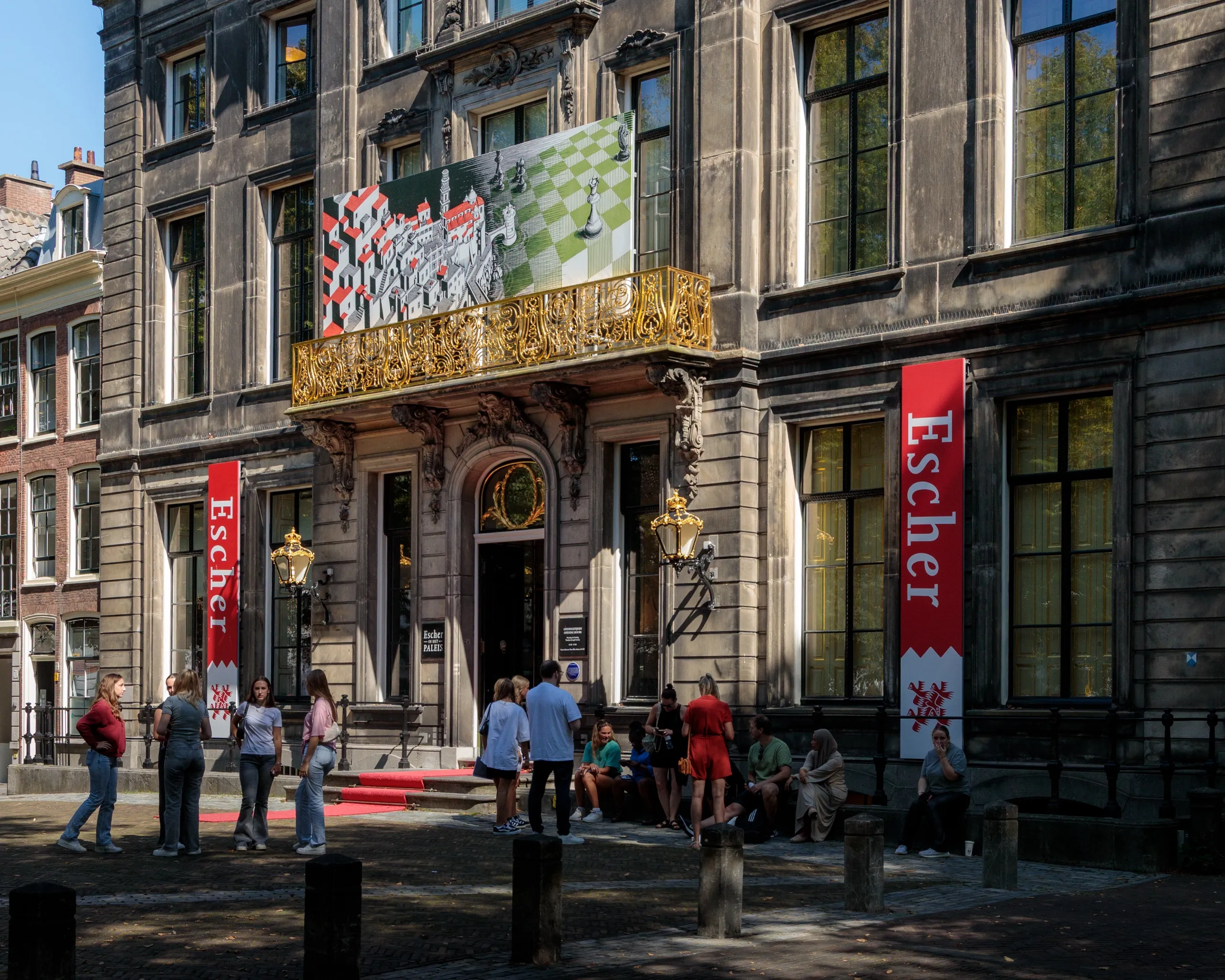 The stone façade of Escher in Het Paleis in The Hague, tall red Escher banners either side of the entrance beneath a gilded balcony.