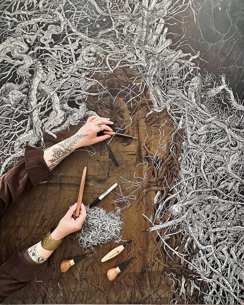 Two hands guiding gouges across the carved lino of Habitat, with cut curls piled at the centre of the block.